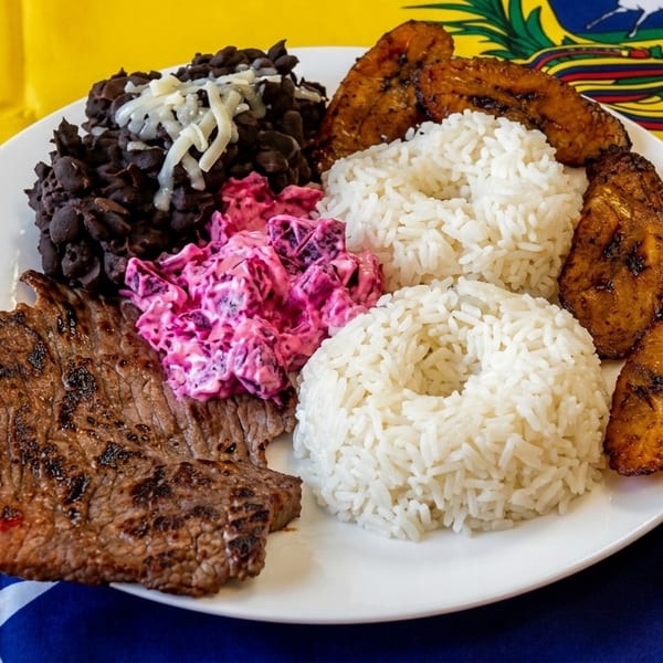Venezuelan meal with grilled steak, black beans, rice, fried plantains, and pink salad on a flag.