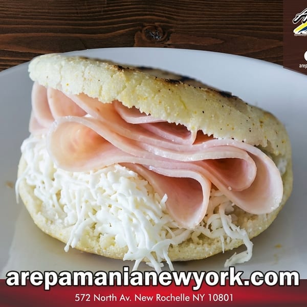 A golden arepa filled with thinly sliced ham and shredded white cheese on a white plate.